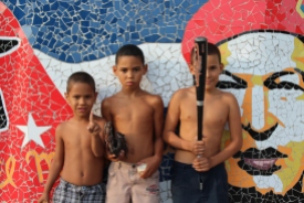Cuban youth hold a donated baseball set in Havana's Fusterlandia neighborhood in April 2016. They are pictured in front of a mosaic mural of Venezuelan leader and Cuban ally Hugo Chavez. The equipment was given to them by a group of Vermont baseball players who visited the island nation.